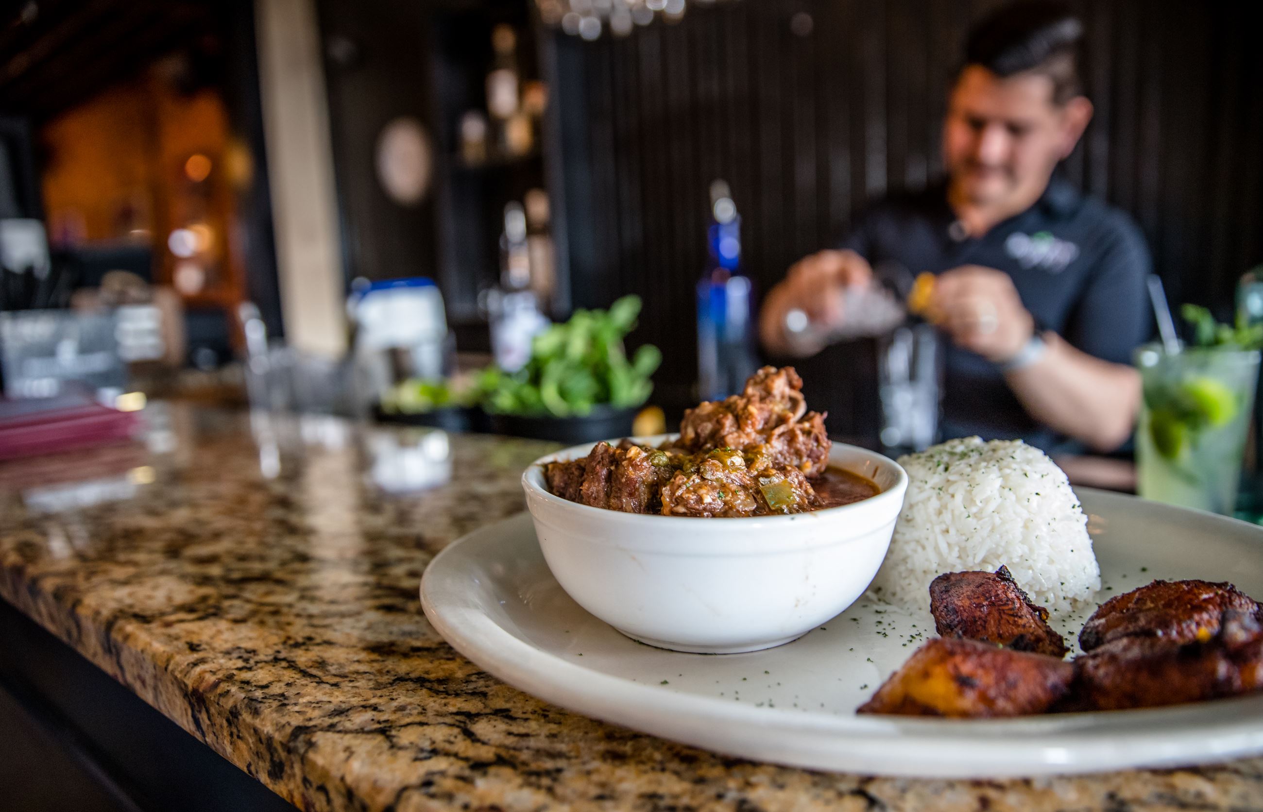 cuban food on counter in front of restaurant bar with bartender mixing drinks in background